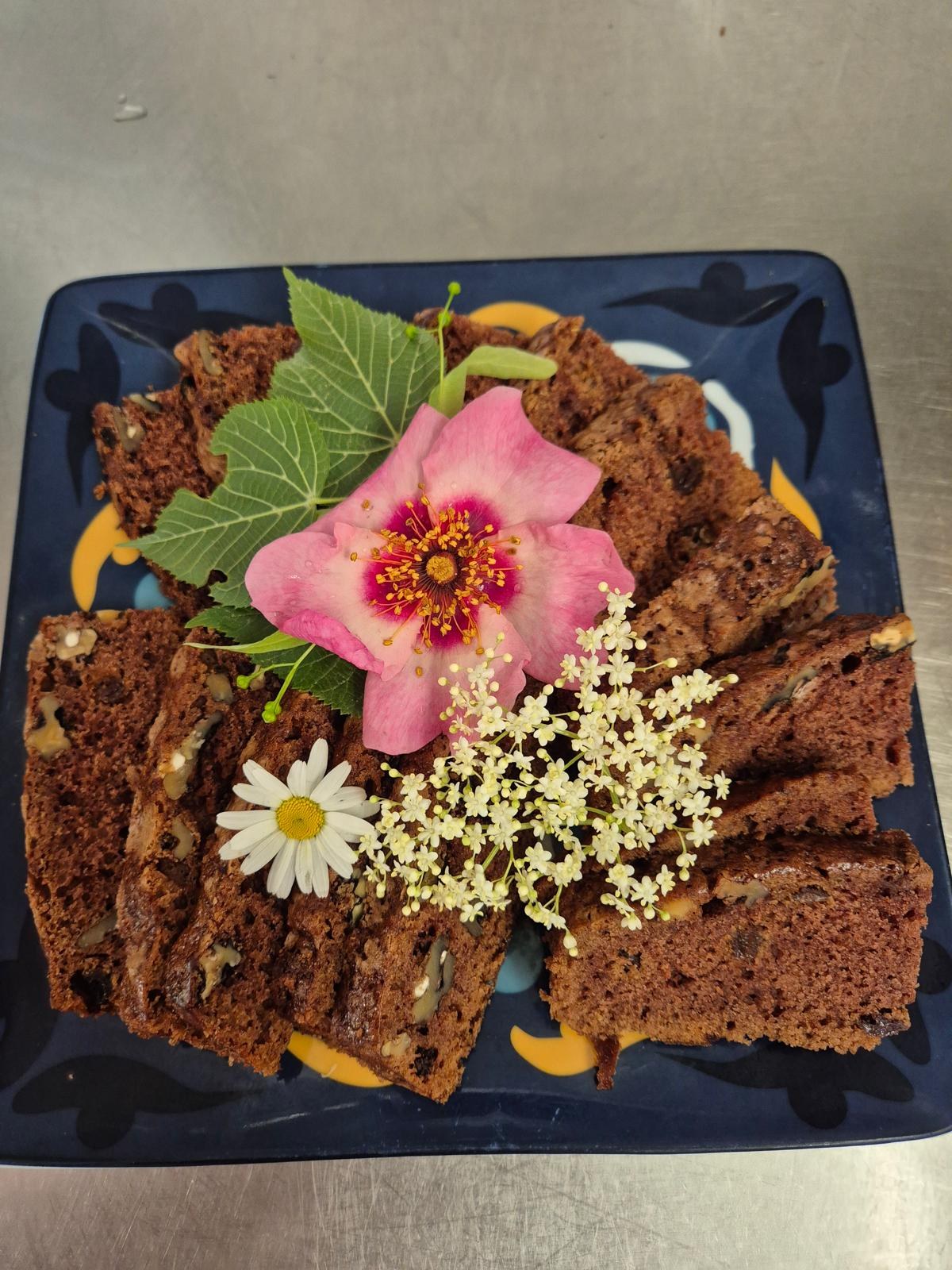 Des parts de gâteau aux noix sur une assiette bleue, décorées d’une rose, d’une marguerite et de fleurs de sureau.