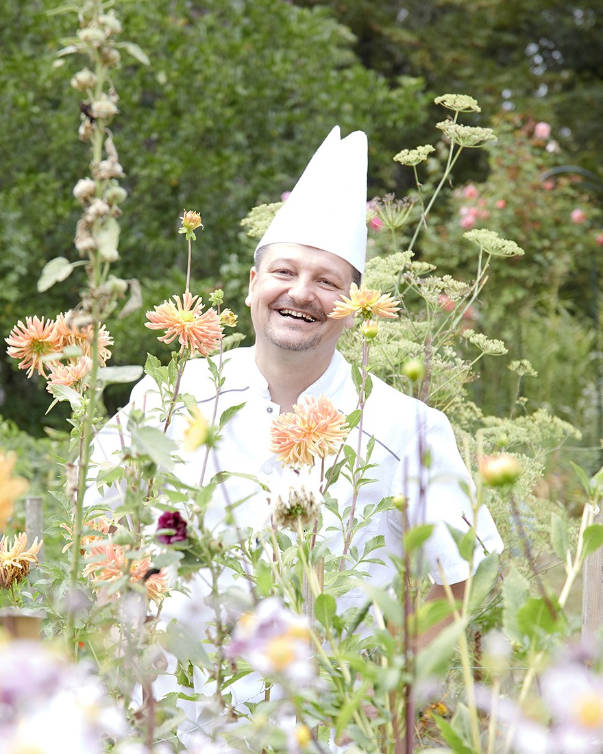 Régis Gagnadre en veste et toque de cuisinier, riant au milieu des dahlias du jardin.